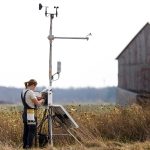 A long shot of a woman standing, working in an electrical box. The box is attached to a tall metal tripod shaped machine. Behind her is another woman with clippers walking towards the the electrical box. They stand in a field with a barn in the background.
