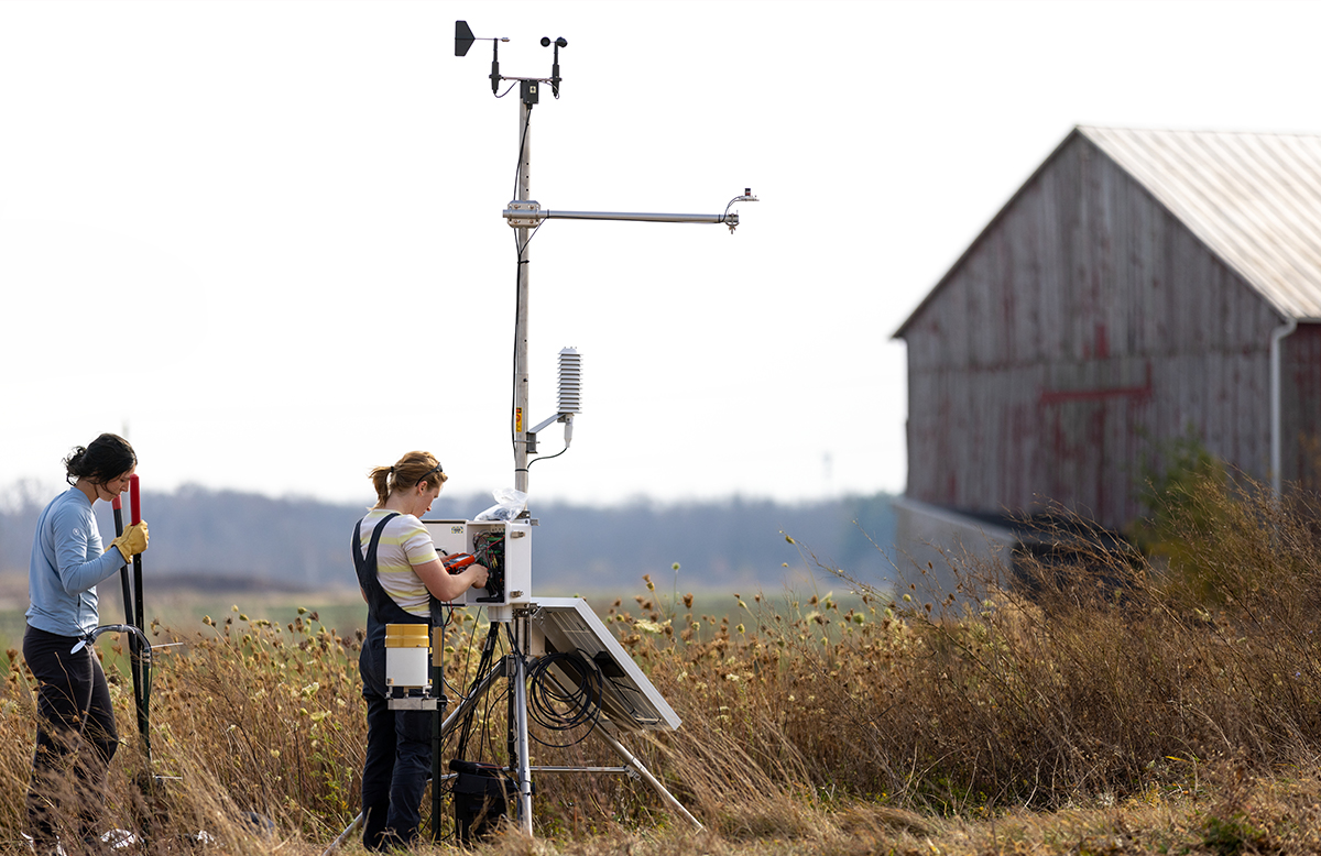 A long shot of a woman standing, working in an electrical box. The box is attached to a tall metal tripod shaped machine. Behind her is another woman with clippers walking towards the the electrical box. They stand in a field with a barn in the background.