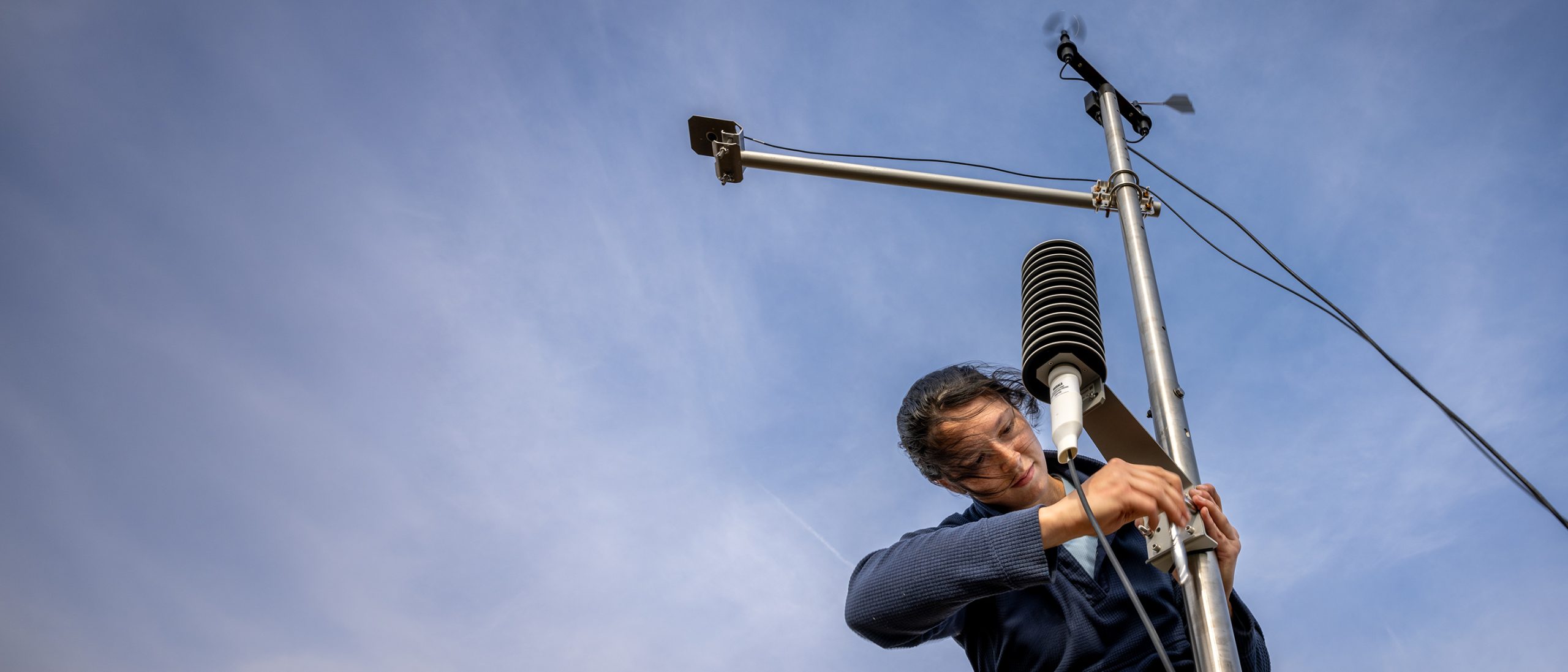 Viewed from below, a woman attaches a piece of equipment to a vertical metal pole, which has a horizontal metal pole and additional equipment attached above her. It's a weather and soil monitoring station. A blue sky in the background takes up the entire frame. The woman's dark hair blows in the breeze.